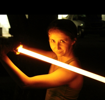 A woman holding a bright orange lightsaber that illuminates her face, arms and upper torso.